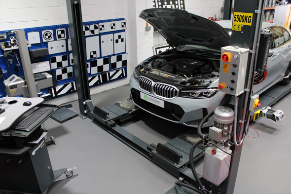 A silver car with an open hood is raised on a car lift in a workshop, surrounded by diagnostic tools and checkered wall panels, showcasing modern automotive technology and maintenance equipment.