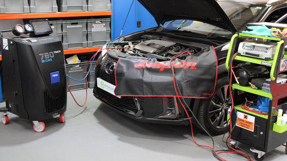 Car undergoing maintenance with an open hood, featuring a Snap-on work mat, diagnostic equipment, and an air conditioning refrigerant machine.