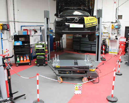 A car lifted on a vehicle hoist in a workshop, surrounded by various automotive tools and equipment, with safety barriers and warning signs indicating electric vehicle precautions.