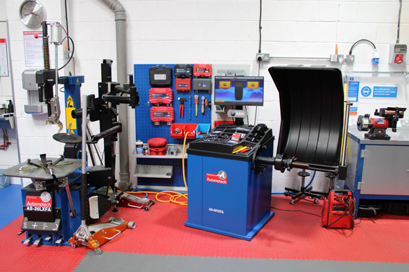 Automotive workshop featuring a tire balancing machine, various tools on a blue wall, and additional equipment, showcasing a well-organized space for tire maintenance and repair.